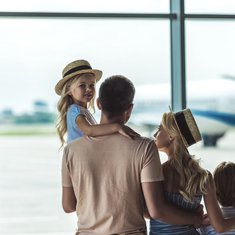 Family with two young children looking out of an airport terminal window at a plane on the tarmac, ready to depart on a holiday