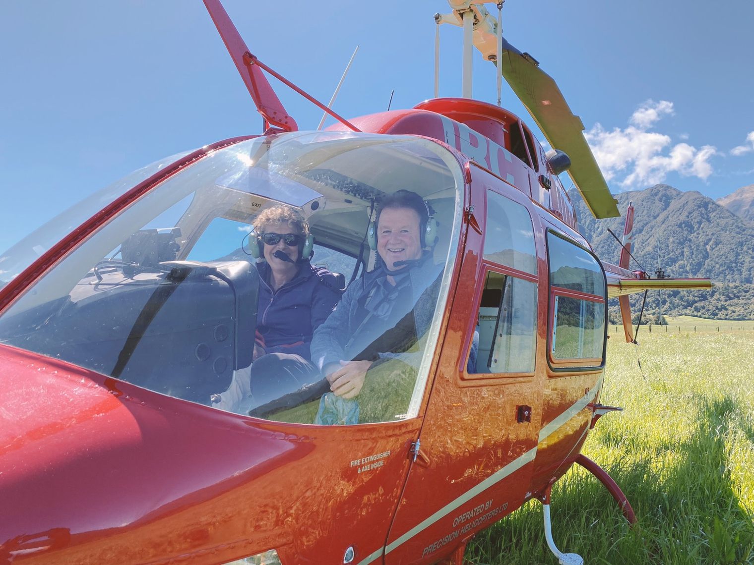 Een felrode helikopter die op een grasveld staat, met passagiers aan boord en bergen op de achtergrond