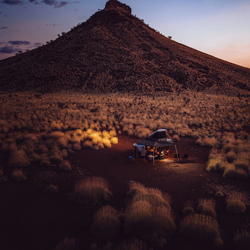 Een camper met een daktent en luifel, warmgloeiend in de schemering in de Australische outback, klein afgezet tegen de iconische piramidevormige Pyramid Hill die uittorent boven rood woestijnstruikgewas onder een diepblauwe avondlucht, Northern Territory, Australië.
