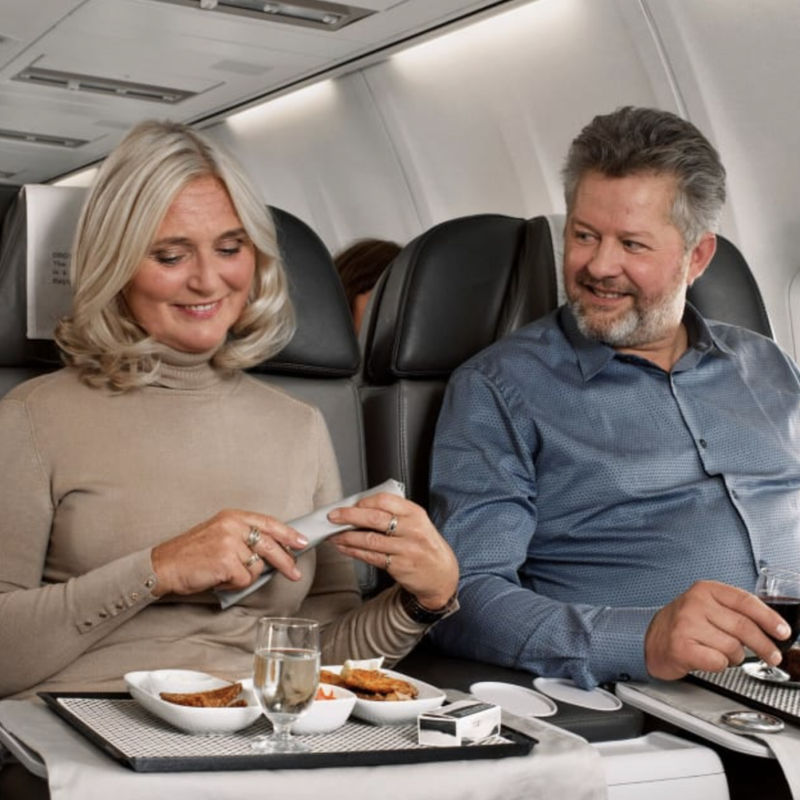 Couple enjoying a meal in premium economy seats on a long-haul flight, with a flight attendant serving in the background