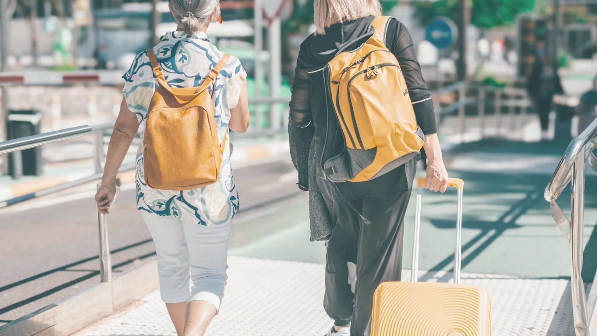 Two people walking to an airport wearing backpacks and one has a pull along suitcase