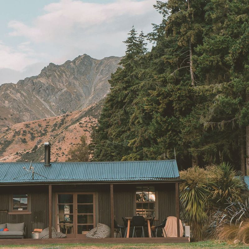Rustiek boerderijhuisje van golfplaat met een overdekte veranda en zitgelegenheid buiten, gelegen tegen de achtergrond van indrukwekkende rotsachtige bergtoppen nabij Lake Hawea, Wanaka, Nieuw-Zeeland