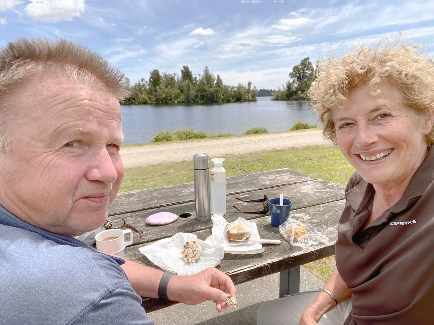 Twee mensen die glimlachend aan een picknicktafel in de open lucht zitten, naast een meer