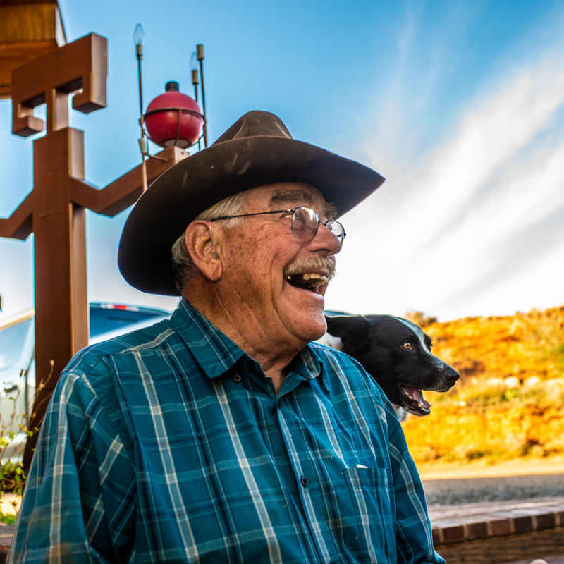 Retired cowboy and guest ranch owner Jerry Kinknade looks to the right of the camera and smiles, his dog and ranch are behind him