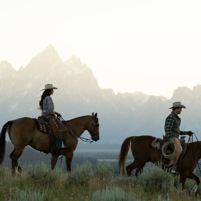 A man and a woman ride horses with a background of mountains in Wyoming, USA