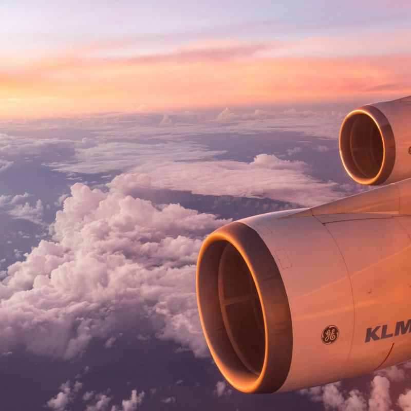 Flügel und Triebwerke eines KLM-Flugzeugs leuchten im weichen Licht eines Sonnenaufgangs über einer Wolkenschicht.