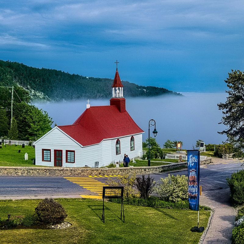 Eine kleine weiße Kirche mit rotem Dach mit Blick auf eine neblige Bucht in Kanada, umgeben von grünen Wiesen und bewaldeten Hügeln.