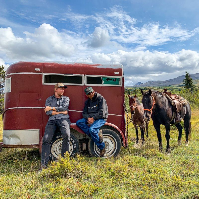 Zwei Männer lehnen an einem roten Anhänger, an dem zwei Pferde angebunden sind, vor dem Hintergrund sanfter Hügel und blauem Himmel in Montana