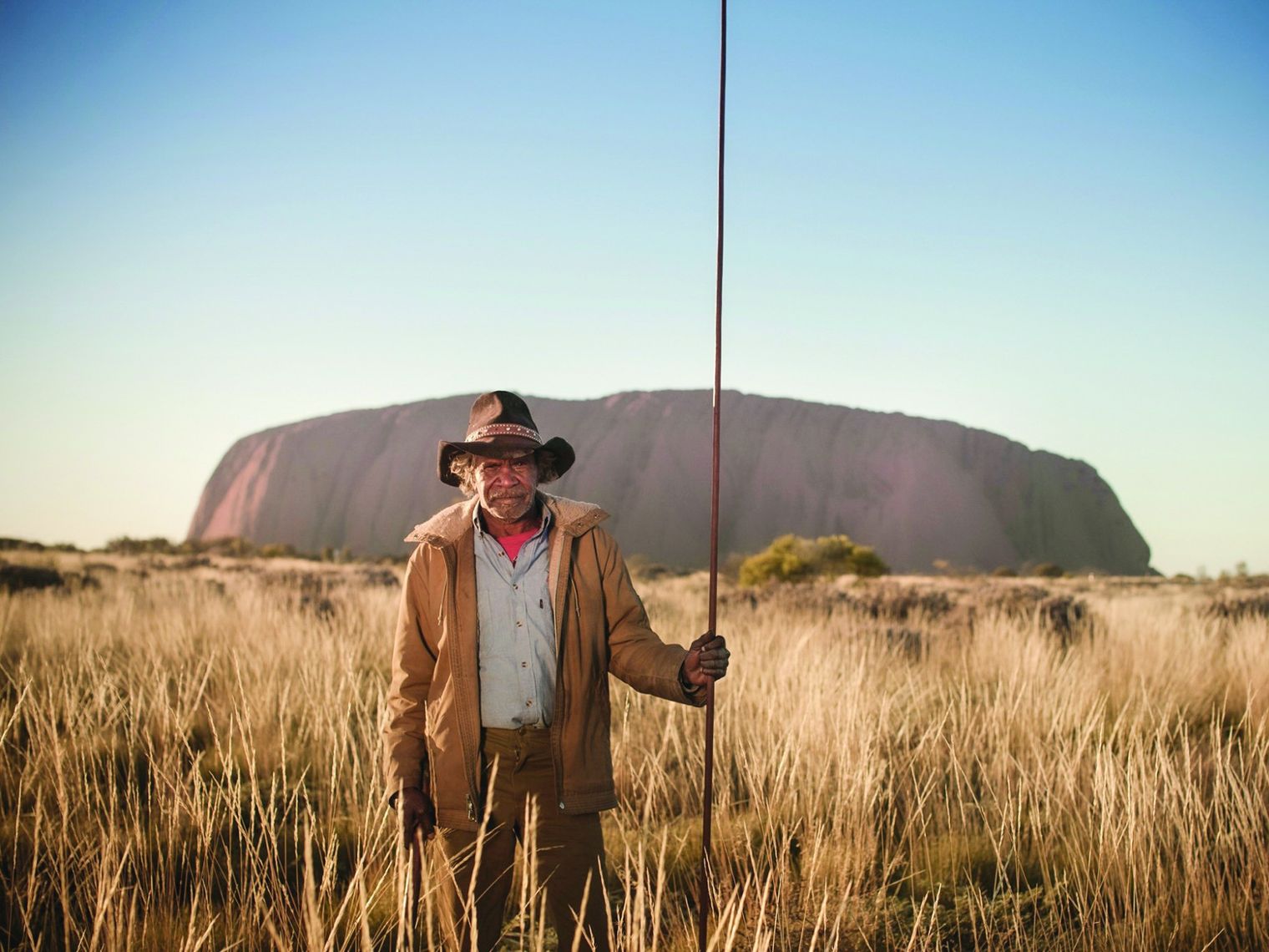 A local guide in Uluru National Park Australia standing in tall grass, holding a staff, with Uluru rock formation in the background.