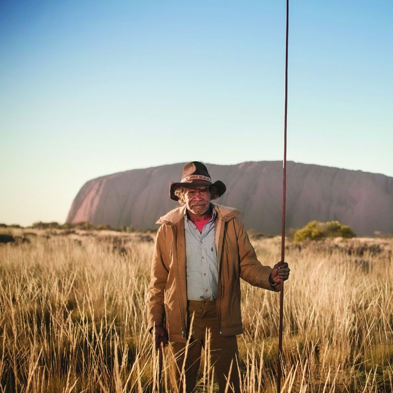 Ein einheimischer Fremdenführer im australischen Uluru-Nationalpark, der im hohen Gras steht und einen Stab in der Hand hält, mit der Felsformation Uluru im Hintergrund.