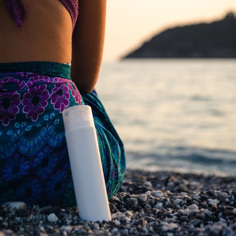 Frau von hinten, die auf Kieselstrand und Gewässer sitzt mit einer Sprayflasche an ihr gelehnt