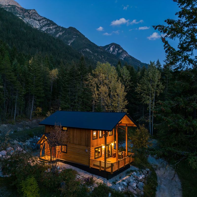 Luftaufnahme einer warm erleuchteten Holzhütte mit überdachter Veranda, eingebettet zwischen Nadel- und Espenbäumen in der Abenddämmerung, mit schroffen Berggipfeln im Hintergrund in Golden, British Columbia, Kanada.