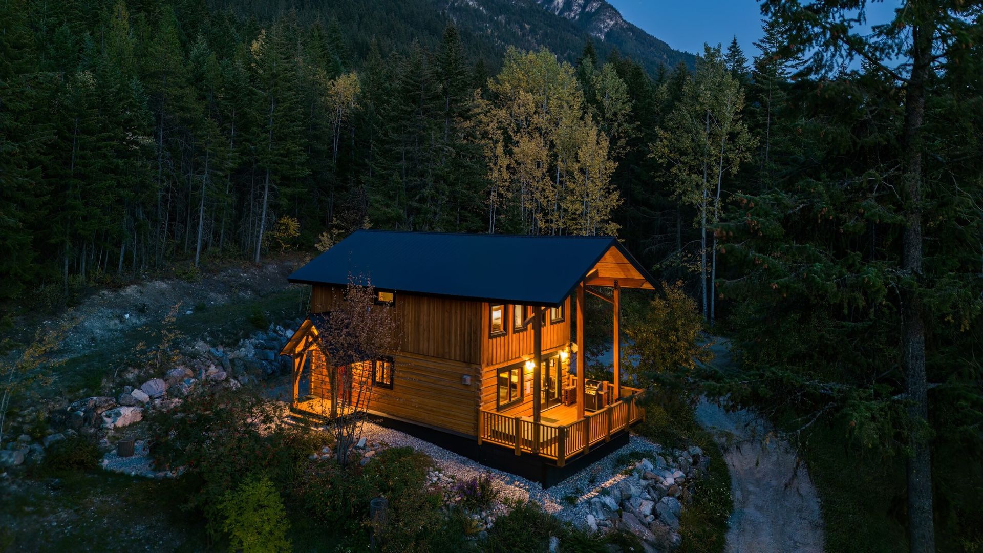 Luftaufnahme einer warm erleuchteten Holzhütte mit überdachter Veranda, eingebettet zwischen Nadel- und Espenbäumen in der Abenddämmerung, mit schroffen Berggipfeln im Hintergrund in Golden, British Columbia, Kanada.