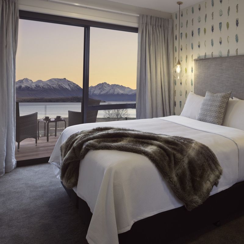 Stijlvolle bed & breakfast-kamer in Nieuw-Zeeland met een kingsize bed, behang met verenprint en een bontplaid, met een eigen balkon met uitzicht op een meer en met sneeuw bedekte bergen bij zonsondergang.