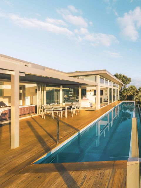 Modernes Glashaus mit Infinity-Pool mit Blick auf grüne Hügel und Wasser bei Sonnenuntergang.