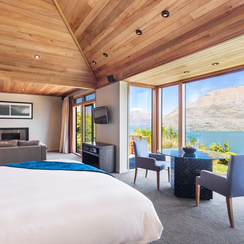 Ruime lodgesuite in Nieuw-Zeeland met een gewelfd houten plafond, kingsize bed, open haard en kamerhoge ramen met uitzicht op een turquoise meer en berglandschap.
