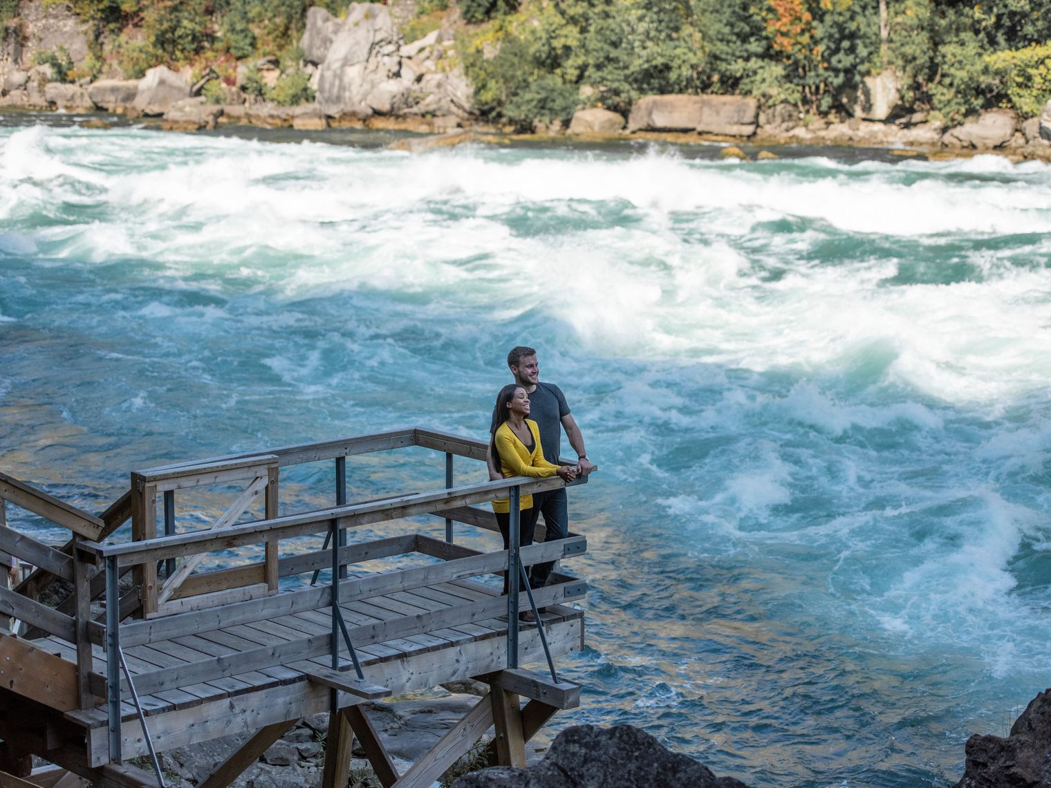 A couple stand on a wooden viewing platform with a fast flowing river running past them at Niagara Falls
