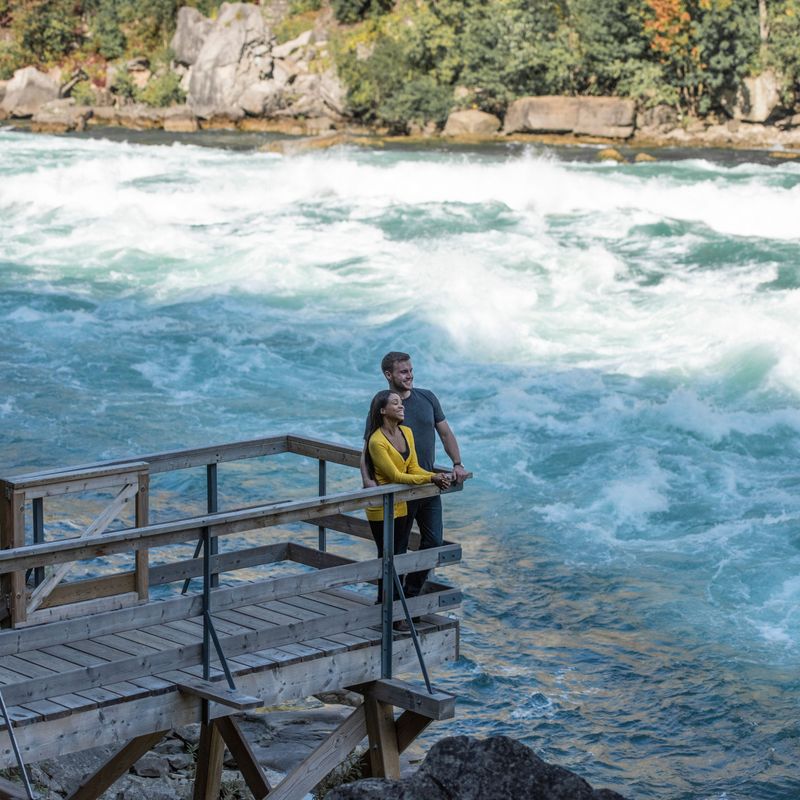 A couple stand on a wooden viewing platform with a fast flowing river running past them at Niagara Falls
