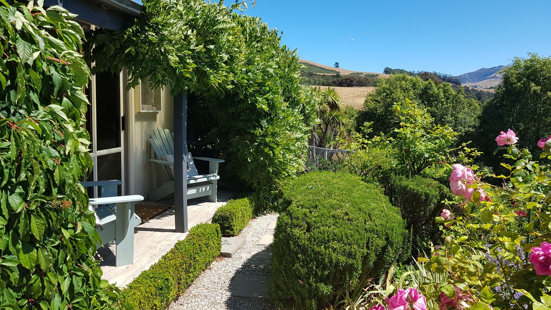 Veranda eines Cottages mit üppigem Garten und sanften Hügeln im Hintergrund, Neuseeland
