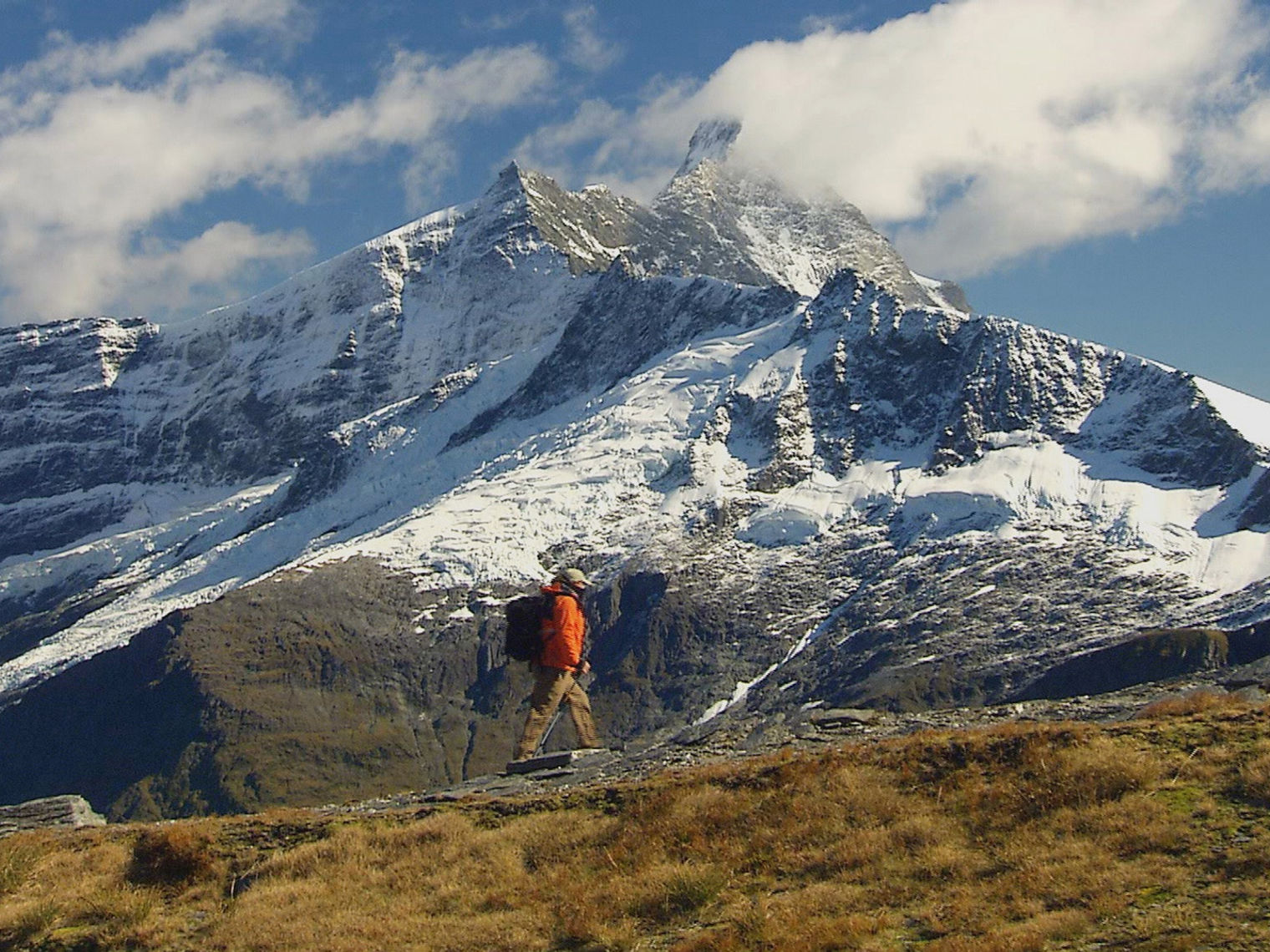 Persoon in een oranje jas loopt door de bergen van Mt. Aspiring National Park met besneeuwde bergtoppen op de achtergrond