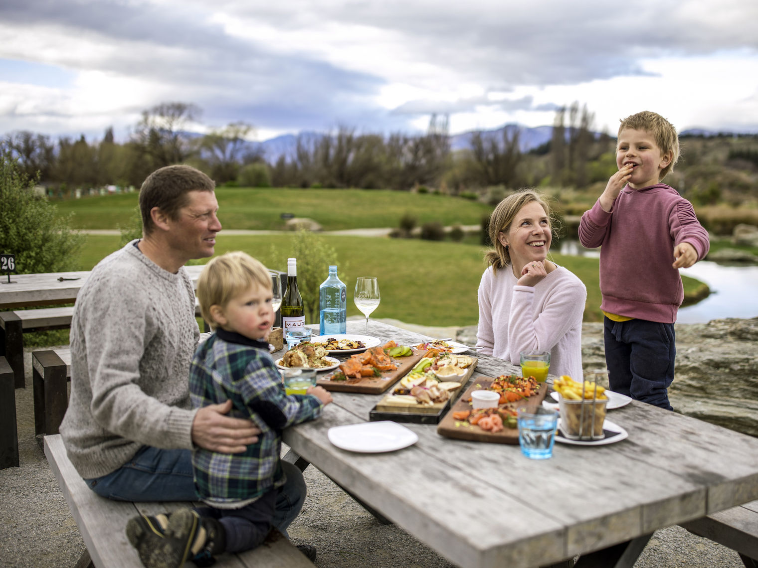 A family sitting down at a picnic table to eat food laid out on wooden boards in Wanaka, New Zealand