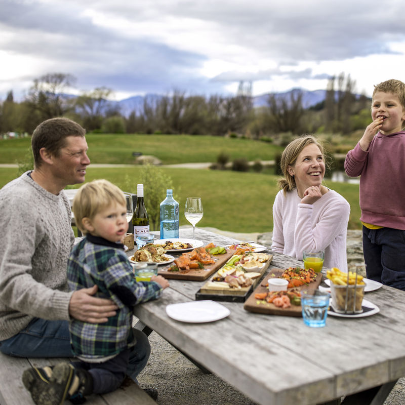 A family sitting down at a picnic table to eat food laid out on wooden boards in Wanaka, New Zealand