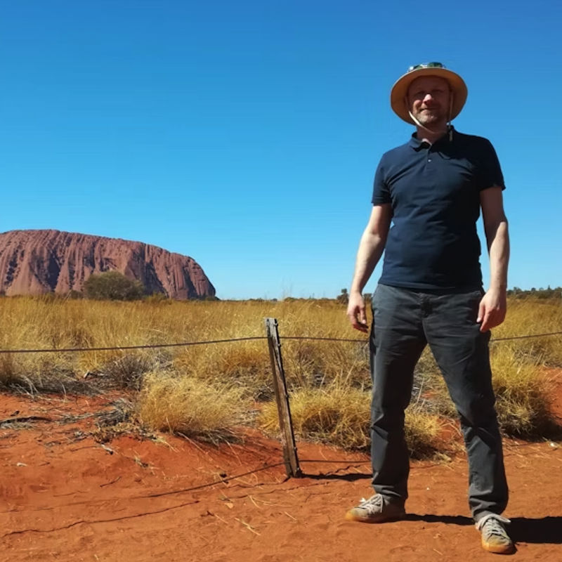 Mann steht auf rotem Sand vor dem Uluru in Australien