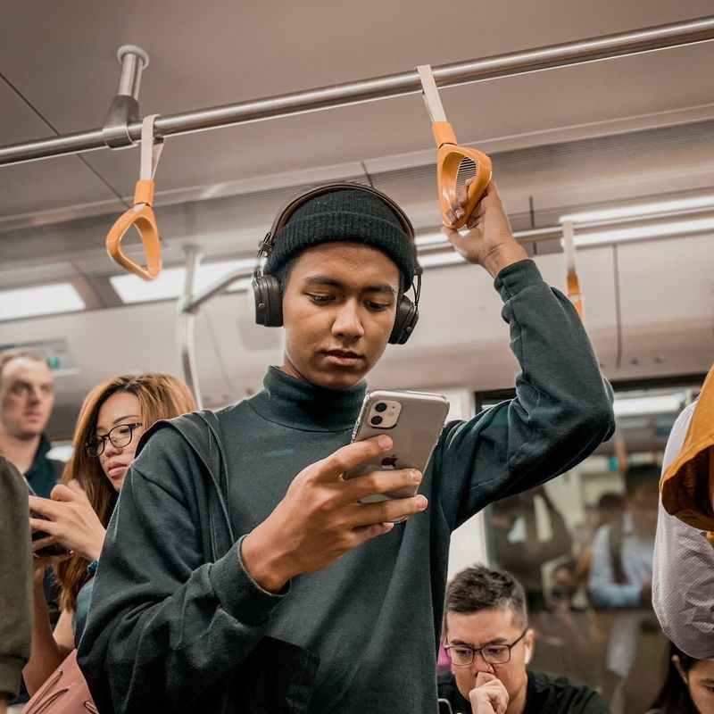 Een man zit in de trein terwijl hij op zijn mobiele telefoon kijkt en een koptelefoon draagt