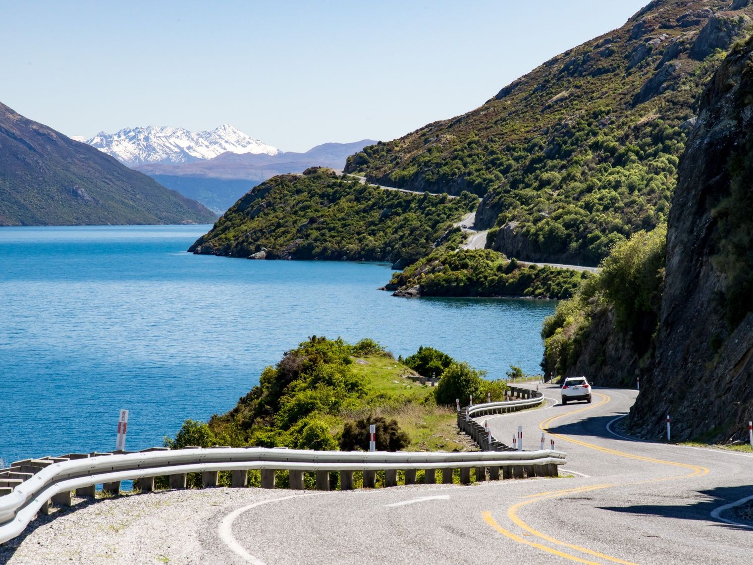 Kronkelweggetje langs Lake Wakatipu waar een witte auto overheen rijdt. In de verte zie je een besneeuwde bergtop en het meer wordt omgeven door groene bergen