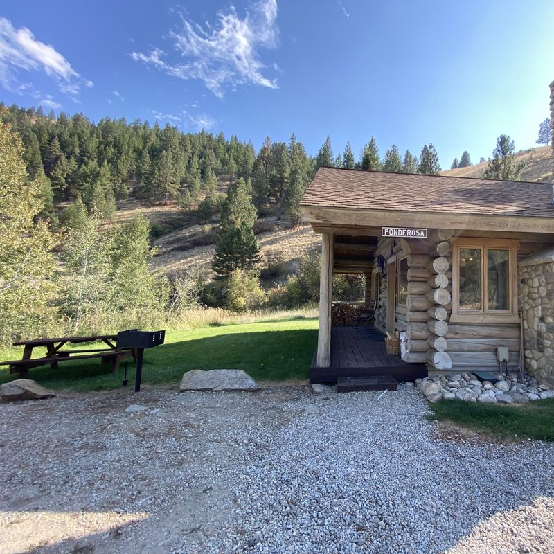 Buitenkant van de Ponderosa blokhut bij Rye Creek Lodge, met een stenen schoorsteen, overdekt terras met gestapeld brandhout en een picknicktafel met BBQ op een grasveld, omgeven door met dennenbomen begroeide heuvels in de Bitterroot Valley, Montana, VS.