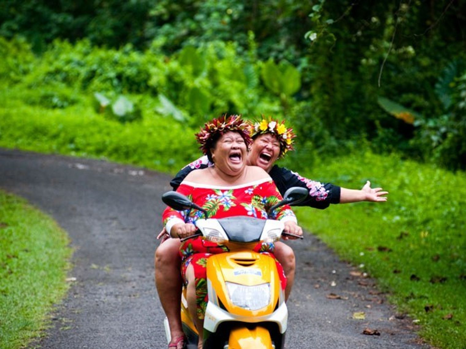 Two Cook Islanders laugh while riding a scooter through the forest
