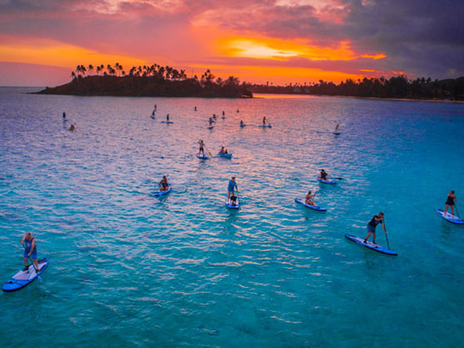 A group of people paddle boarding in the bays of Cook Islands with a colourful sky in the background