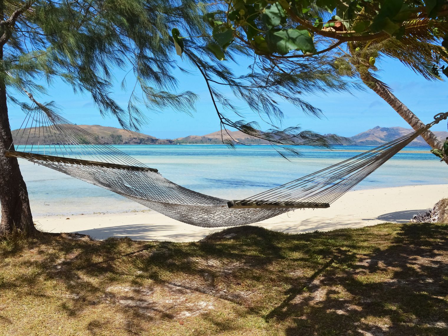 Eine zwischen zwei Bäumen am Strand gespannte Hängematte mit dem Meer und anderen Inseln im Hintergrund