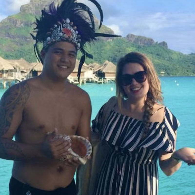Sylvie en een Maori-man lachen naar de camera met de oceaan en bergen op de achtergrond
