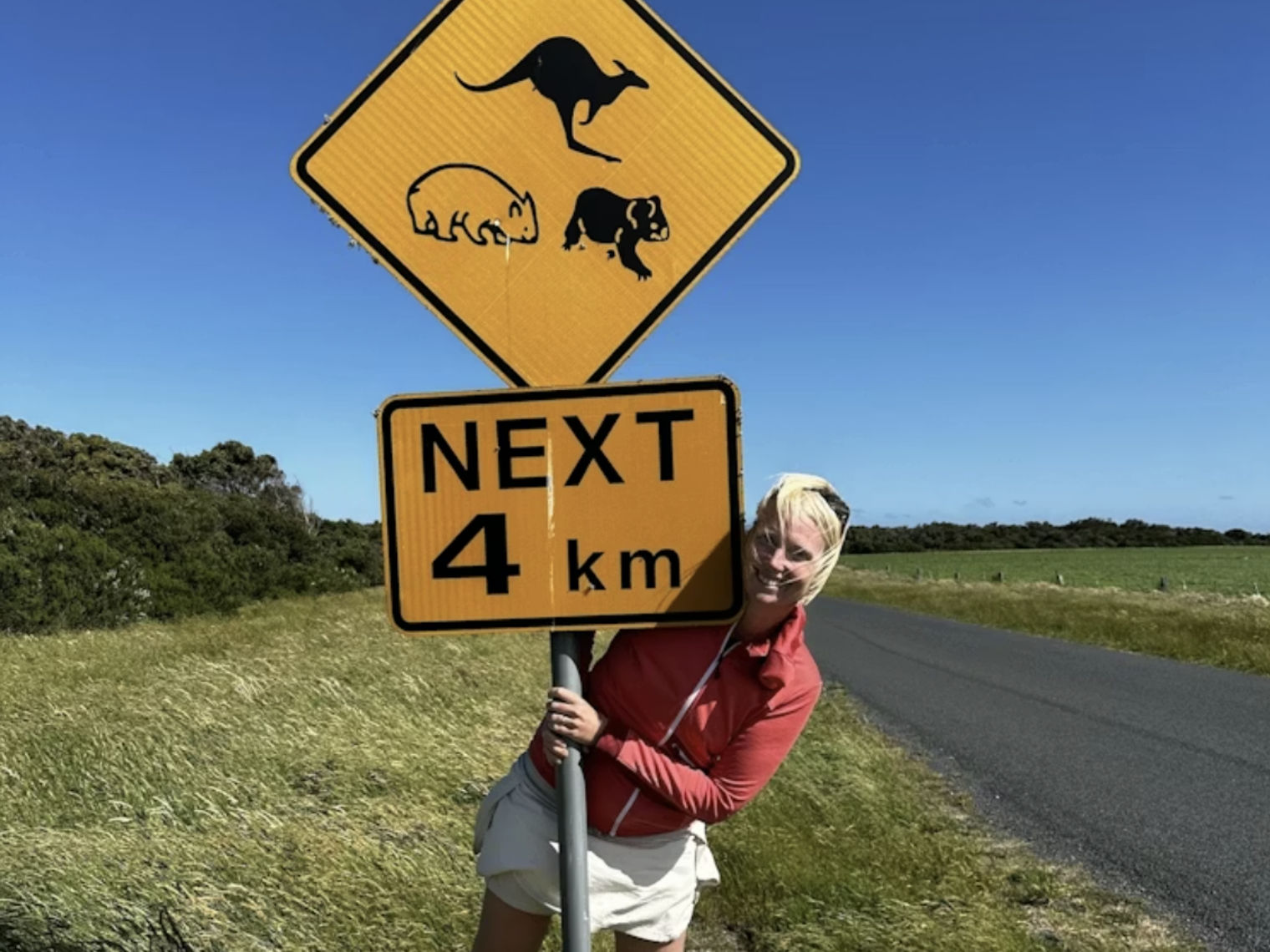 Frau steht neben einer Straße und umfasst ein australisches Verkehrsschild mit Tiersymbolen und Hinweis "Next 4 km"