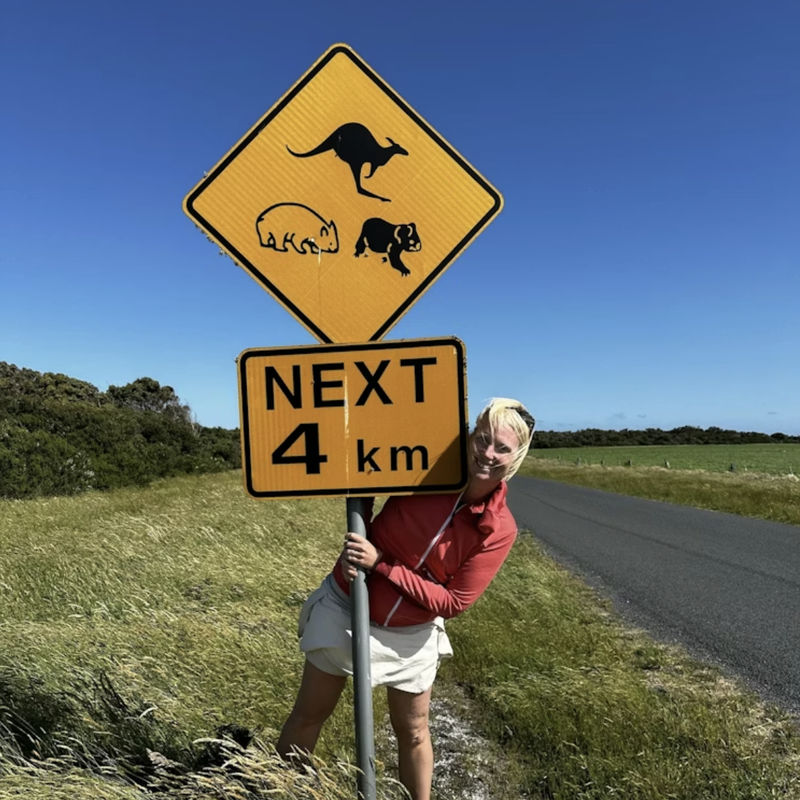 Frau steht neben einer Straße und umfasst ein australisches Verkehrsschild mit Tiersymbolen und Hinweis "Next 4 km"