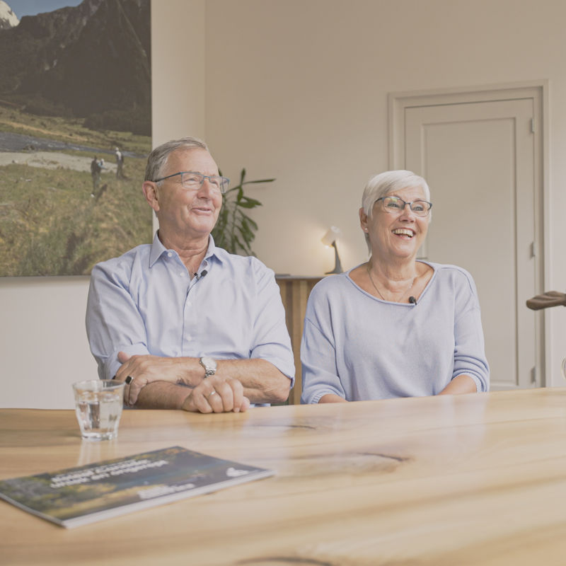 Klanten Job en Nelly aan een houten tafel tijdens een planningsgesprek met hun reisexpert, glimlachend terwijl ze hun aankomende reis bespreken, met een grote foto van een helikopterexcursie in Nieuw-Zeeland aan de muur achter hen.
