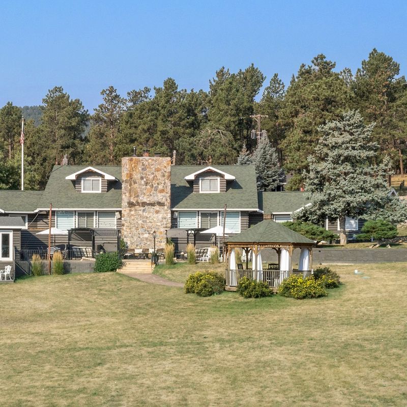 Rustikales Blockhaus-Inn mit hohem Steinkamin und Pavillon auf einer offenen Rasenfläche, umgeben von kiefernbedeckten Hügeln in der Black Hills Region von South Dakota, USA