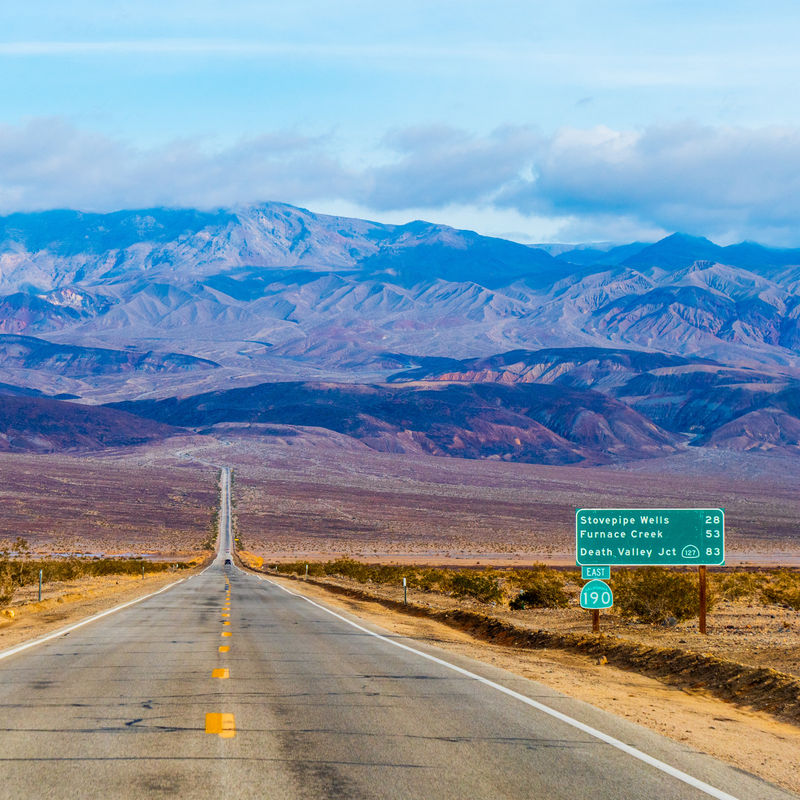 Een lange rechte weg die zich uitstrekt door de Mojavewoestijn van Californië naar de bergen van Death Valley, met woestijnvlakten en bergkammen in de verte onder een heldere hemel.