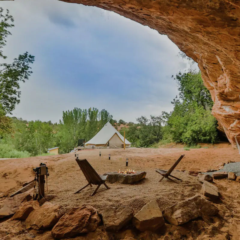 Unieke glampinglocatie onder een natuurlijke zandstenen rotsoverhang in Utah, VS, met een canvas bell-tent, kampvuur en houten stoelen in een rood woestijnlandschap.