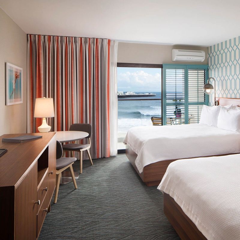 Lichte hotelkamer in retrostijl in Santa Cruz, Californië, VS, met twee queensize bedden, blauwgroen geometrisch behang en een balkon met uitzicht op de oceaan en de pier.