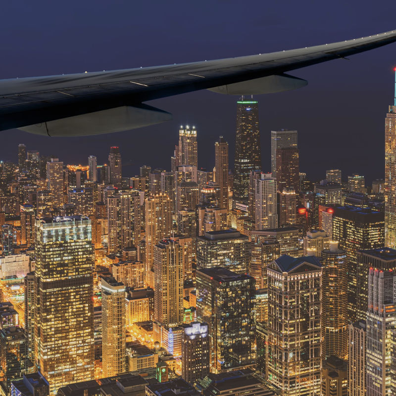 Aerial view of Chicago's illuminated skyline at night from an airplane window, with the city's iconic skyscrapers glowing below the aircraft wing, USA