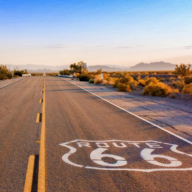 Een leeg stuk Route 66 door de woestijn bij zonsopgang, met lange schaduwen en bergsilhouetten in de verte.