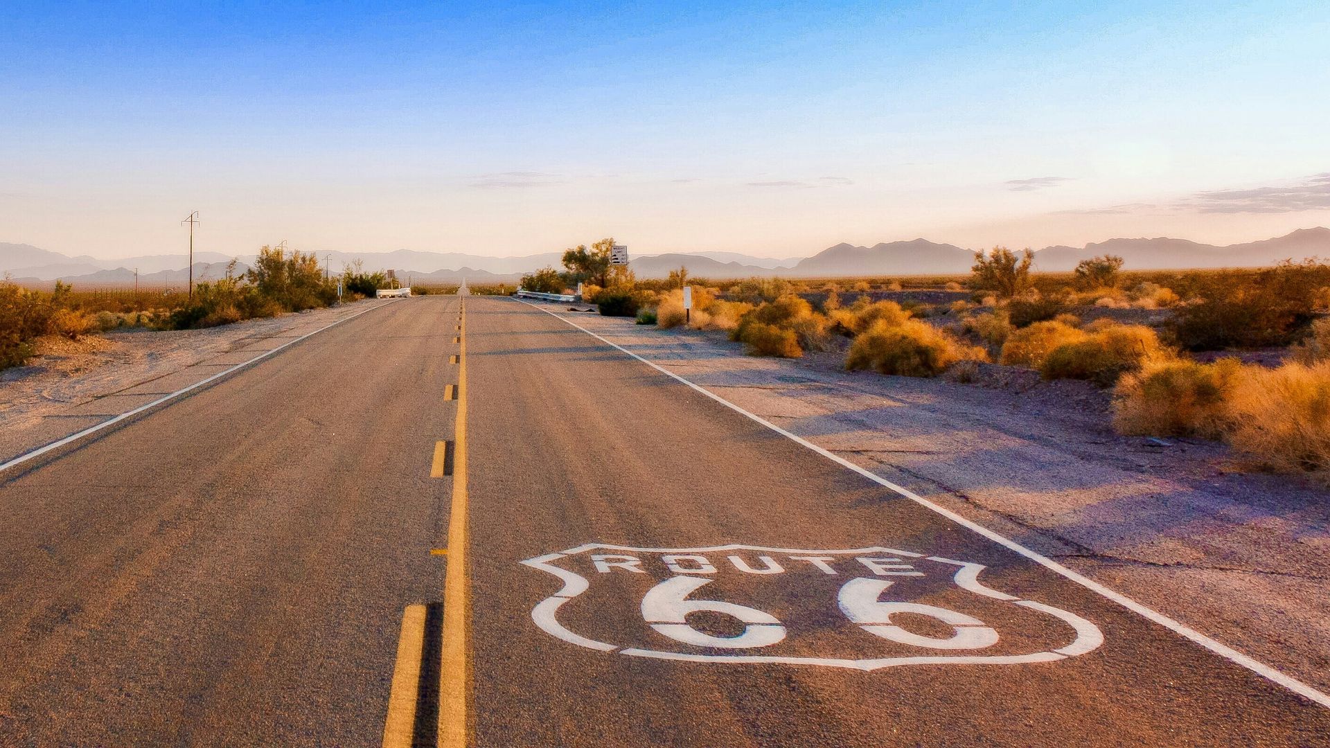 Eine leere Strecke der Route 66, die bei Sonnenaufgang durch die Wüste führt, mit langen Schatten und fernen Bergsilhouetten.