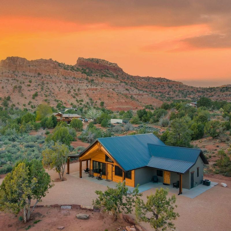 Aerial view of Casa Vermillion, a modern cabin with a teal metal roof and covered porch, set amid desert scrubland with dramatic red rock mesas and a vivid orange sunset sky near Kanab, Utah, USA.