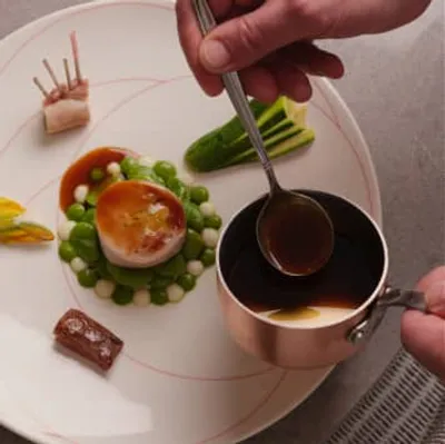 A chef elegantly plates a gourmet dish, delicately drizzling sauce from a copper pan.