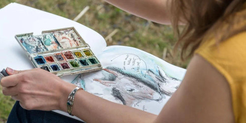 A woman is painting with watercolors on a piece of paper.