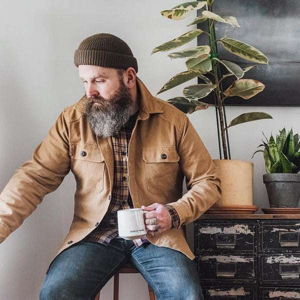 A man sitting on a stool next to a cabinet while drinking tea