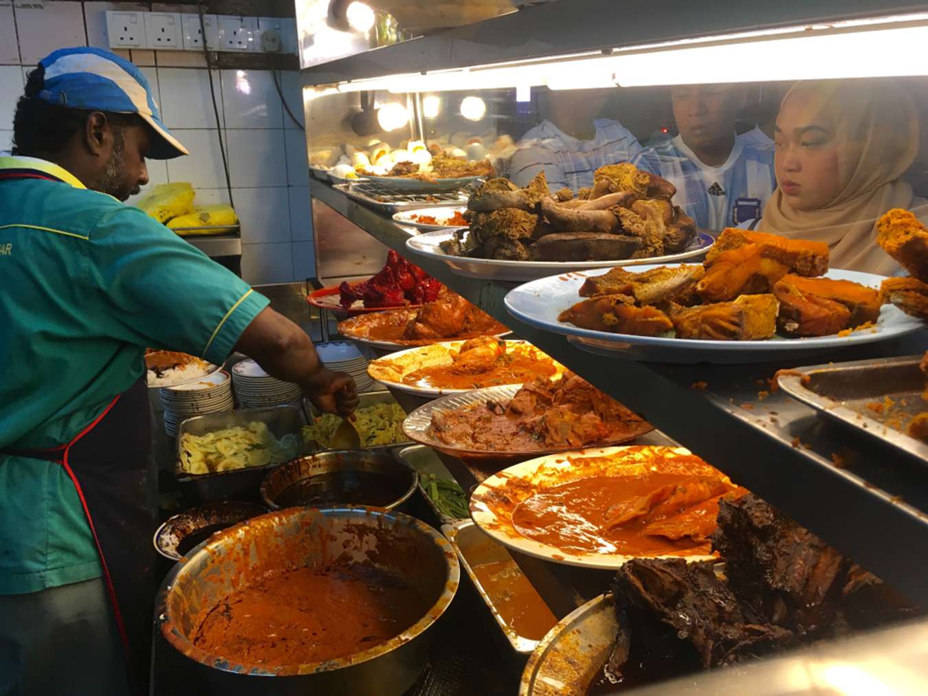 Customers look on the dishes as a worker prepares the meal at a nasi kandar restaurant in Kuala Lumpur,