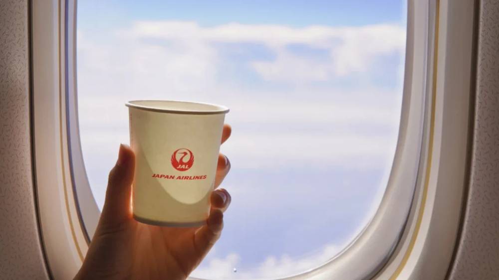 A hand holding a cup in front of a window on a Japan Airlines plane.